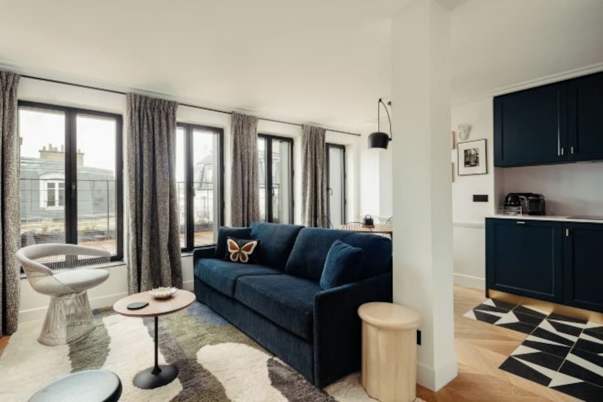 Una sala moderna con sofá, una butaca, una mesita y una zona de cocina con armarios y encimera.