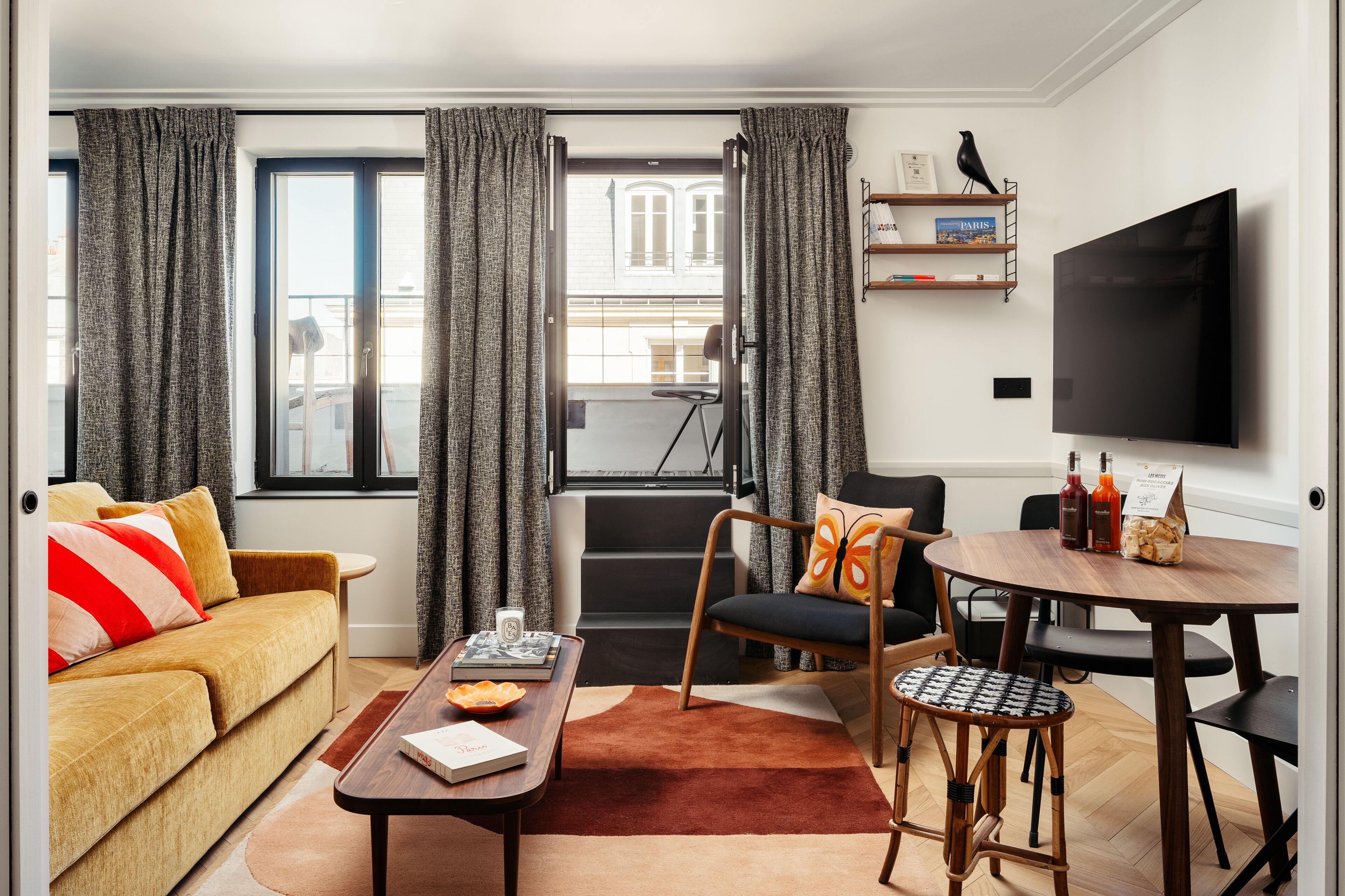 A modern living room with a sofa, coffee table, and a dining area with chairs. There is a TV mounted on the wall and a balcony visible through the window.