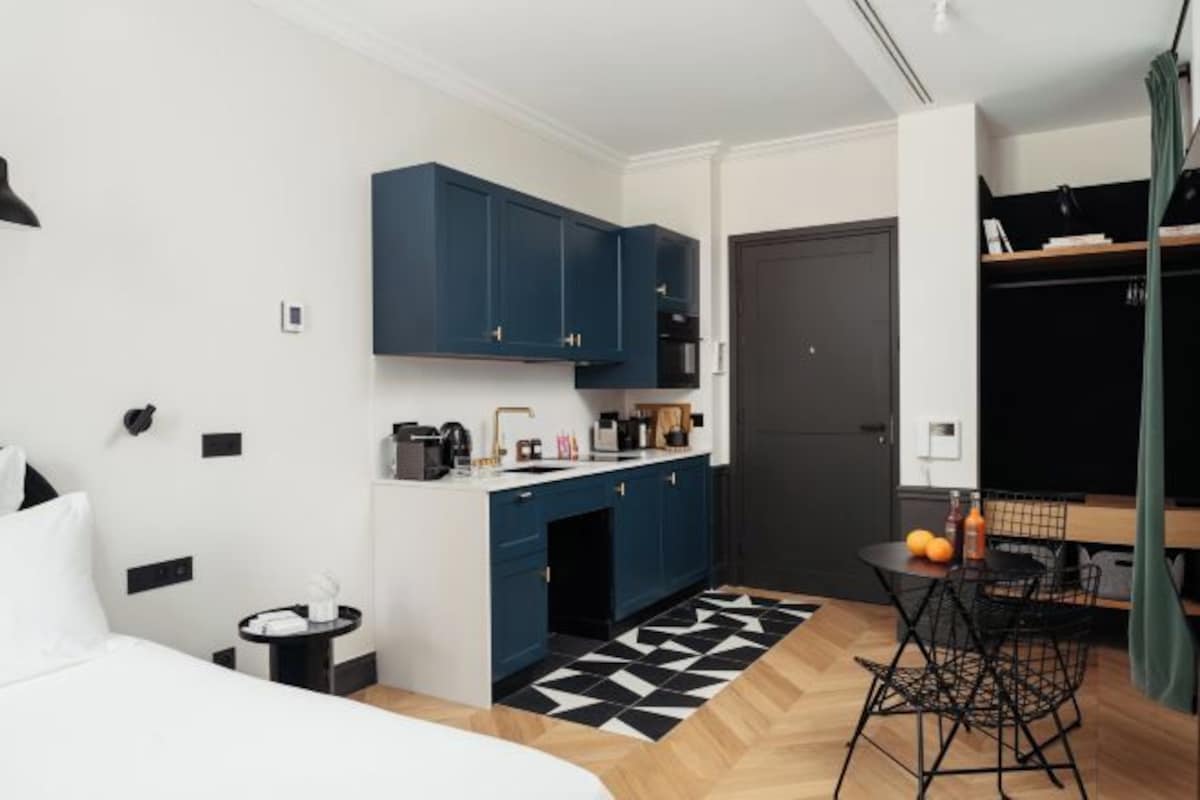 Una habitación de hotel compacta con kitchenette, una cama, una mesita con sillas y una alfombra con estampado blanco y negro.