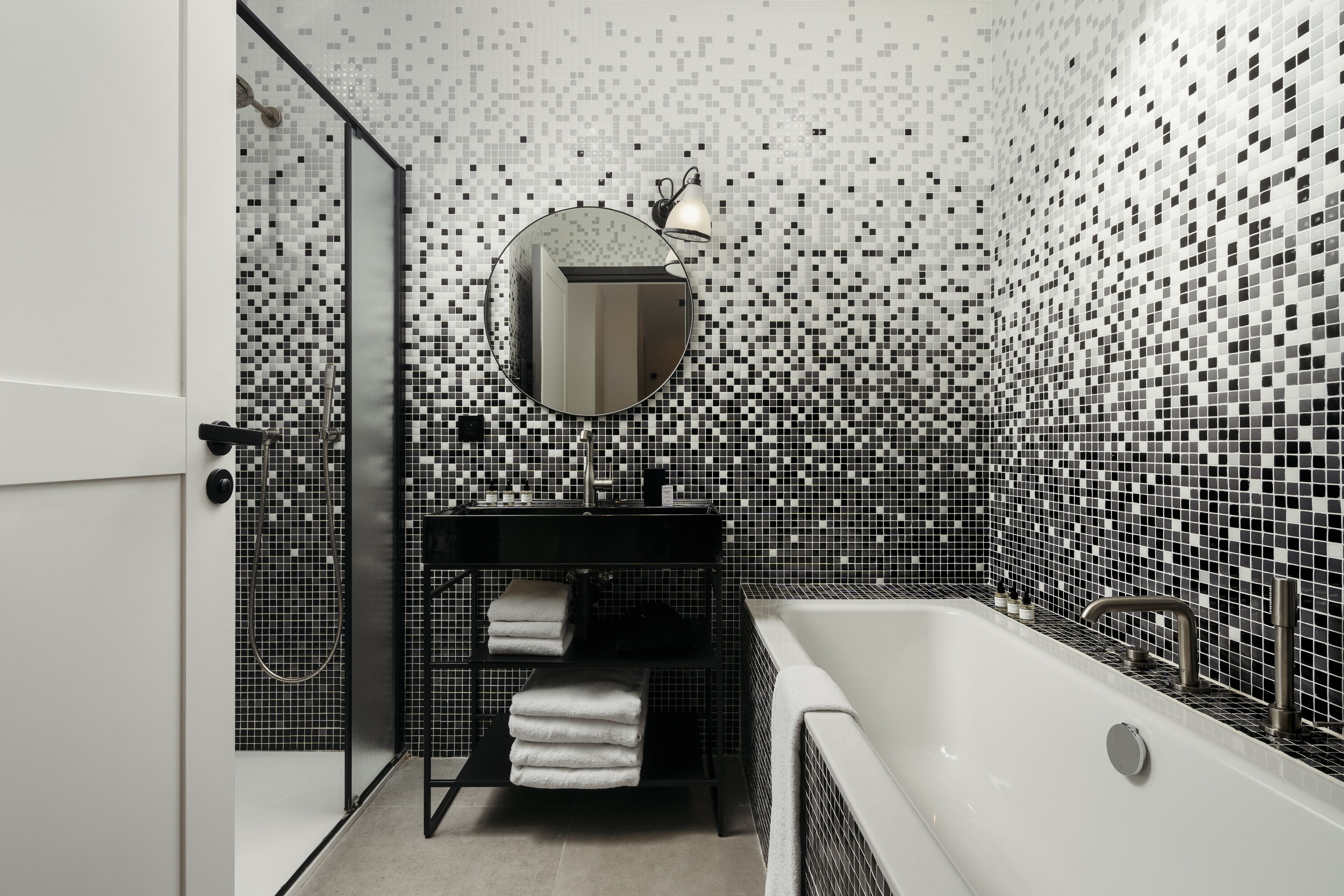 A modern bathroom with a black and white mosaic tile wall, a round mirror, a black shelf with towels, and a white bathtub.