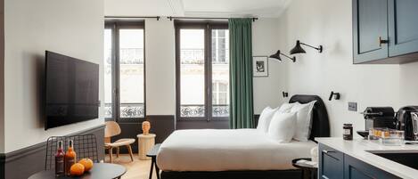 A modern hotel room with a bed, a dining table, a TV, and a kitchenette.