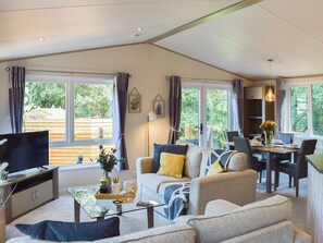 A modern living room with a sofa, coffee table, and TV. There are large windows with curtains, a dining area with chairs, and a kitchen visible through the glass door. - 3 bedroom accommodation in Perthshire (Perthshire)