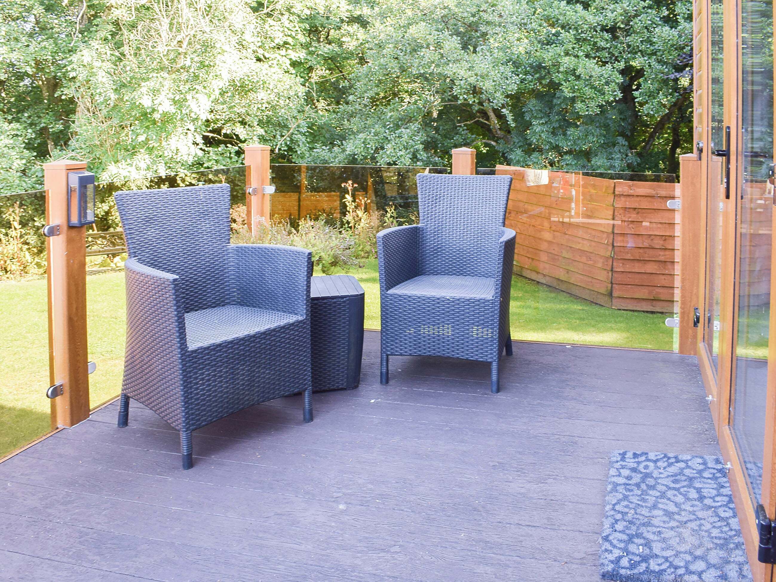 Eine Terrasse mit zwei Korbstühlen und einem kleinen runden Tisch, umgeben von einem Holzzaun und üppiger Bepflanzung.