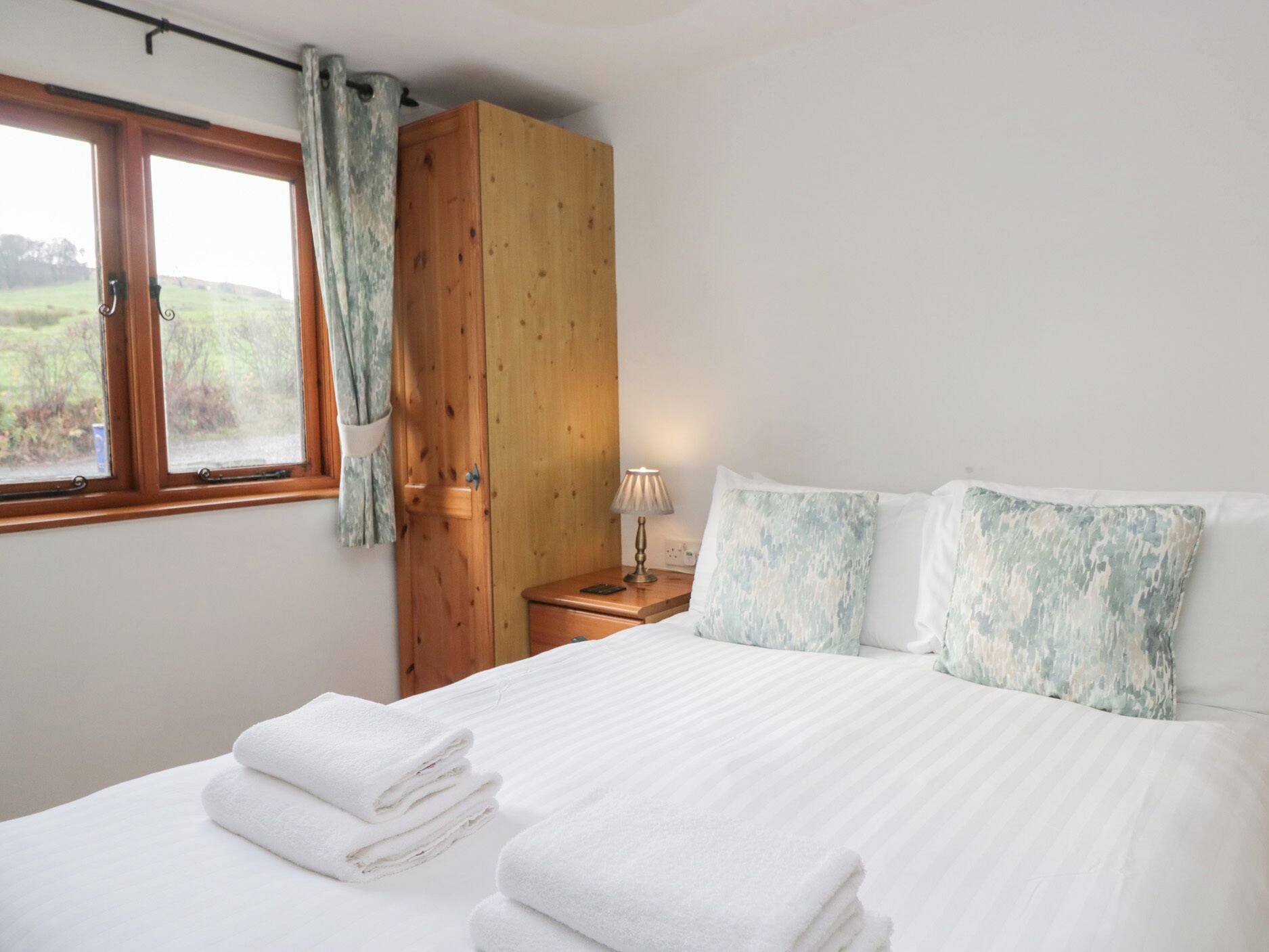 A bedroom with a wooden wardrobe, a bedside table with a lamp, a bed with white linens, and a window with a view of greenery.