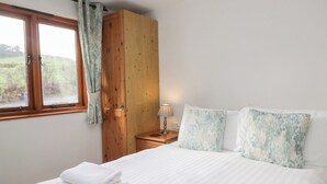 Une chambre avec une armoire en bois, une table de chevet avec une lampe, un lit avec du linge de maison blanc et une fenêtre donnant sur un paysage verdoyant.