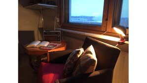 Ein gemütliches Zimmer mit Fenster, Tisch mit Büchern, Sessel mit Kissen und Lampe.