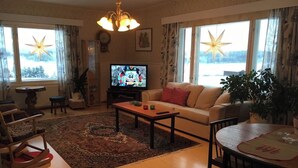 Ein gemütliches Wohnzimmer mit einer Couch, einem Couchtisch und einem Fernseher. Große Fenster mit Vorhängen, eine Kristallleuchter und eine Uhr an der Wand sind ebenfalls vorhanden.