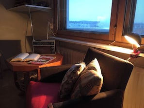 Une pièce confortable avec une fenêtre, une table avec des livres, une chaise avec un coussin et une lampe.