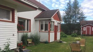 Ein Haus mit einer kleinen Veranda, ein Garten mit trittfesten Steinen und ein rotes Bauernhaus im Hintergrund.