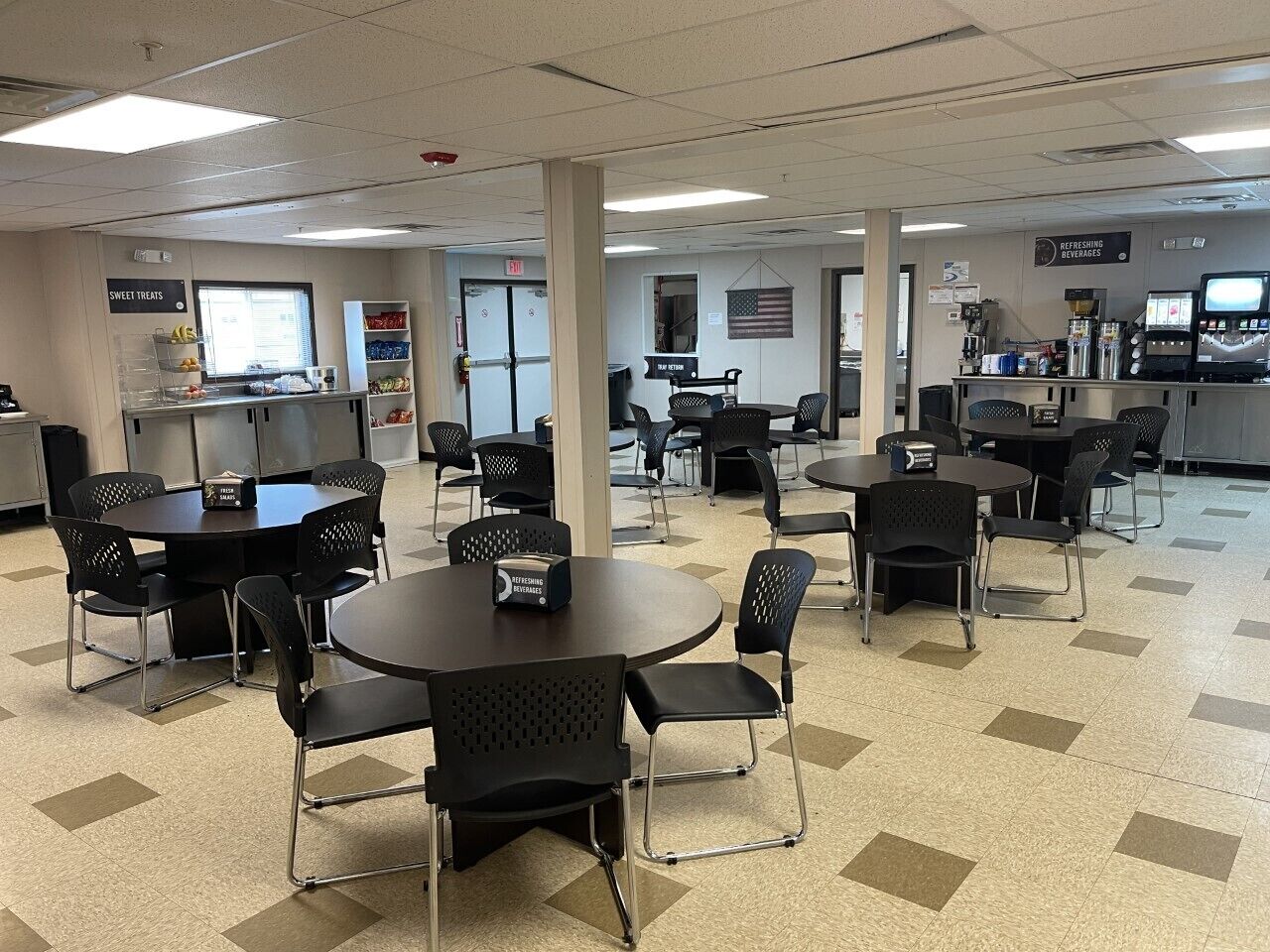 A cafeteria with multiple round tables and chairs, a counter with a sign for sweet treats, and a refreshment area with a coffee machine.