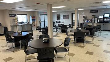 A cafeteria with multiple round tables and chairs, a counter with a sign for sweet treats, and a refreshment area with a coffee machine.