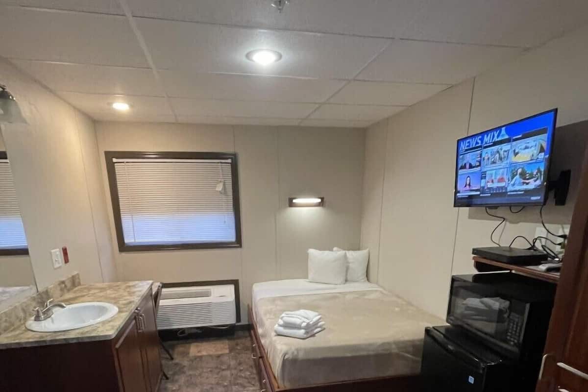 Habitación de hotel compacta con cama, un pequeño escritorio, televisor, microondas y baño con lavabo y espejo.