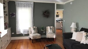 Ett vardagsrum med en soffa, två fåtöljer, ett soffbord och en matta. Det finns ett fönster med gardiner, en väggdekoration och ett kök i bakgrunden.