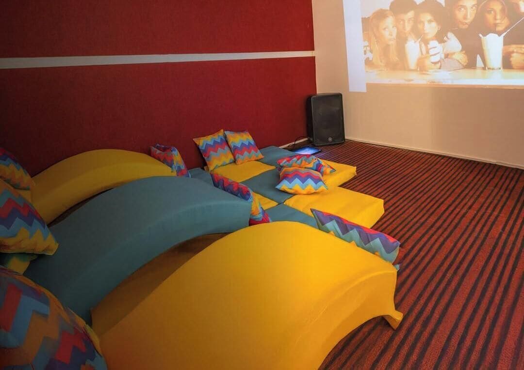 Una stanza con un grande schermo che mostra 'Friends', posti a sedere colorati e una parete di accento rossa.