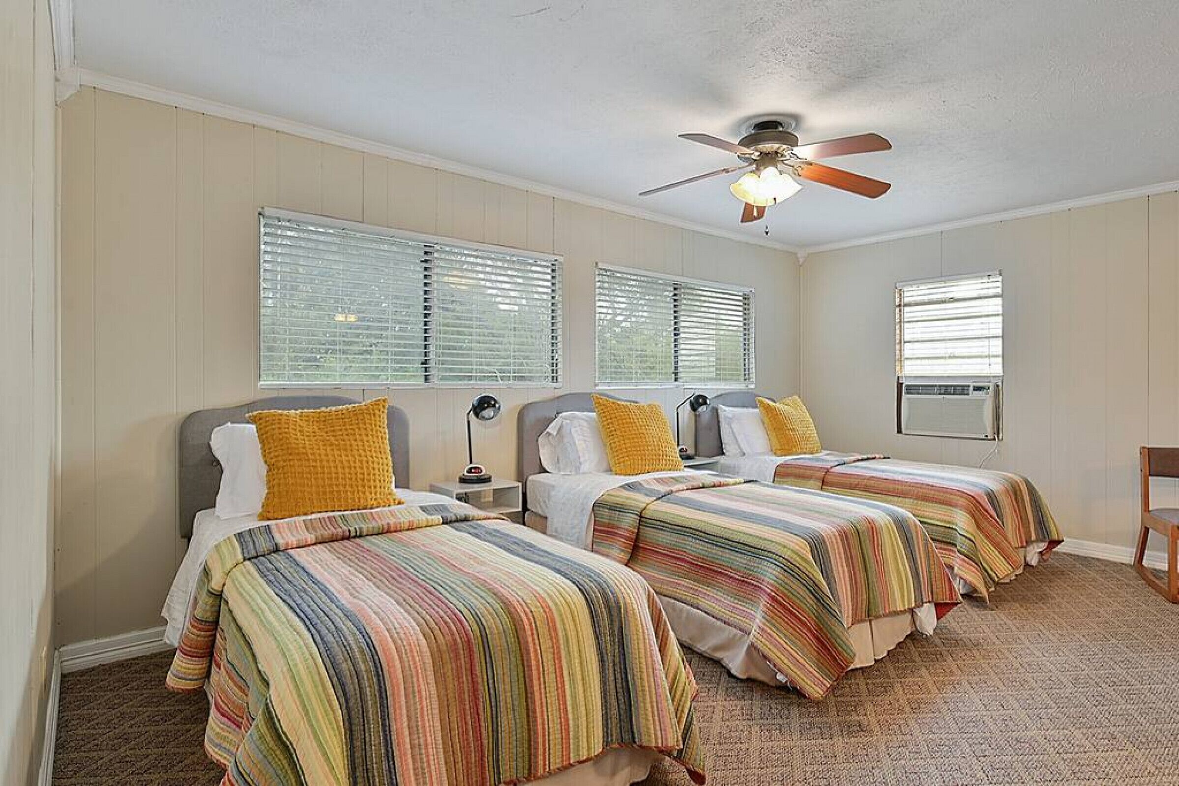 A hotel room with three beds, a ceiling fan, and large windows with blinds.
