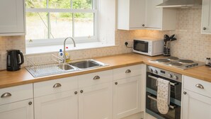 A modern kitchen with white cabinets, a stainless steel sink, a microwave, a stove, and a window with a view of greenery outside. - UK35715 - South Cottage (Howpasley, near Hawick)
