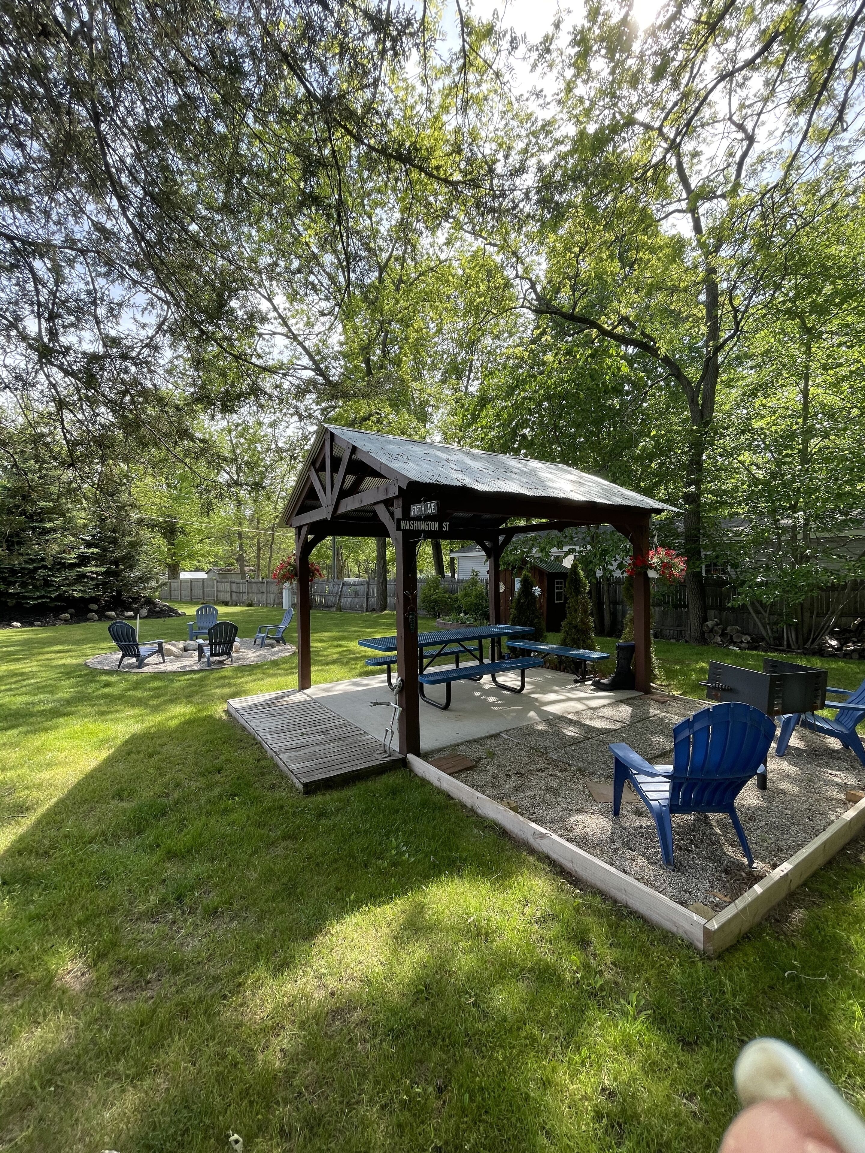A covered picnic area with benches and chairs, surrounded by a grassy lawn and trees.