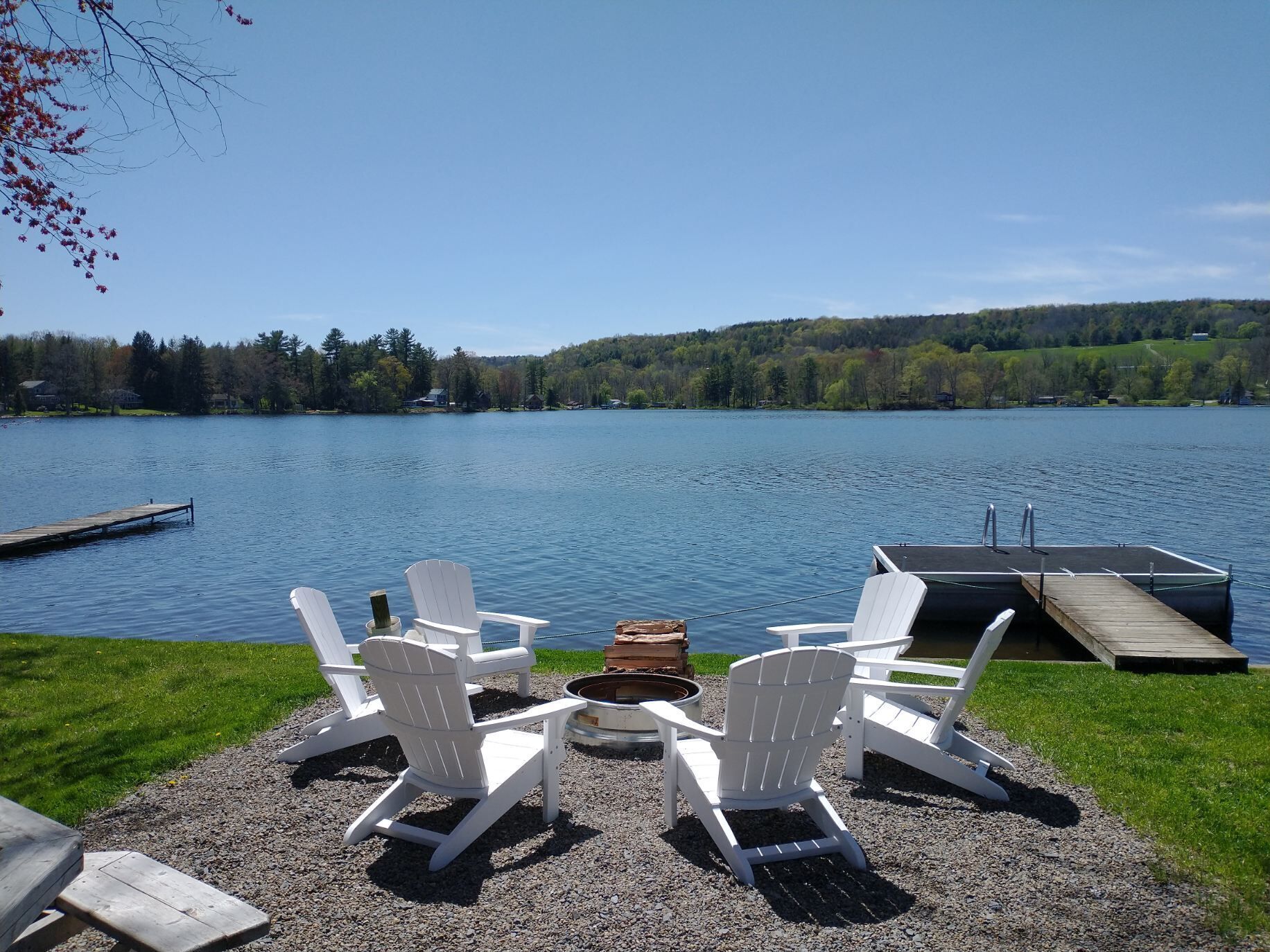 A lakeside view with a dock, three Adirondack chairs, and a small fire pit.