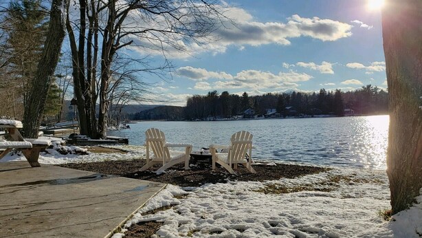 Una terrassa al costat del llac amb cadires Adirondack i una taula de picnic, tot envoltat de terreny i arbres coberts de neu.