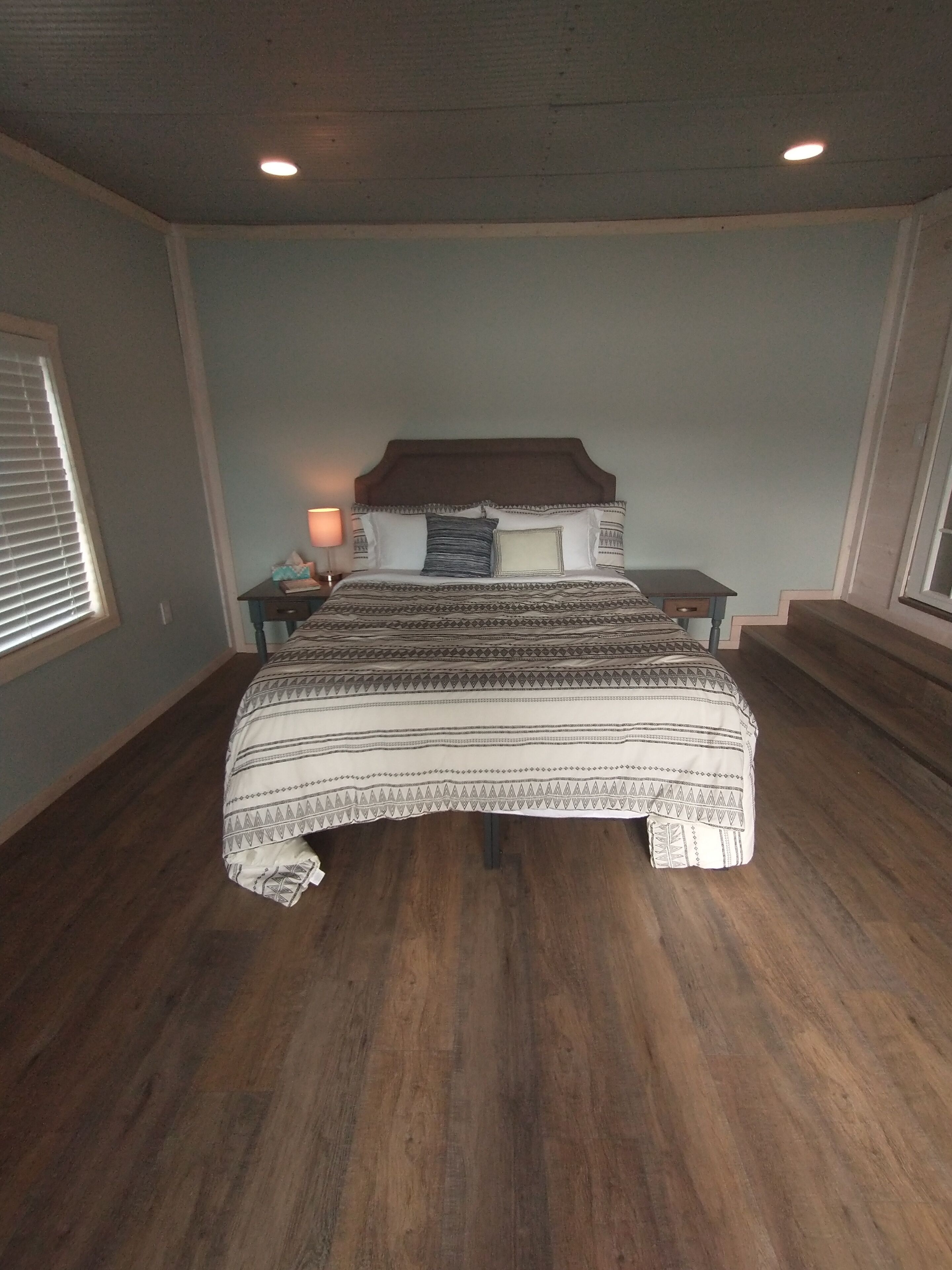 A bedroom with a large bed, wooden flooring, a nightstand with a lamp, and a window with blinds.