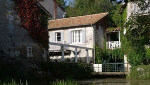 A rustic building with a tiled roof, a balcony, and a bridge over a calm water body.