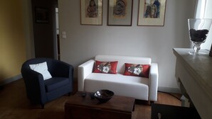 A living room with a dark blue armchair, a white sofa with red floral cushions, a coffee table, and a small bowl. There are framed artworks on the wall and a glass of coffee on the windowsill.