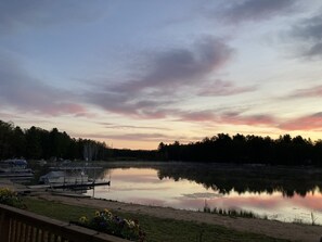 Ein ruhiger See im Abendlicht, mit am Ufer links verankerten Booten, umgeben von Bäumen und einer blumigen Wiese.