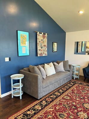 Ein Wohnzimmer mit blauer Wand, grauem Sofa, Teppich mit Muster und gerahmten Kunstwerken.