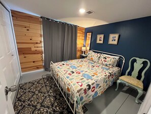 Ein Schlafzimmer mit einem Bett, einem Sessel, einem gemusterten Teppich, Holzpaneelen an der Wand, einem grauen Vorhang und zwei gerahmten Bildern.