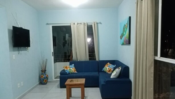 A living room with a blue sofa, a small wooden table, a TV mounted on the wall, and a window with curtains.