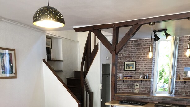A cozy living area with a wooden dining table, two wooden chairs, a pink sofa, and a staircase leading to a kitchen area with a brick wall and bar stools. - House with garden and free parking, 2 minutes from Le Clos Lucé (Amboise)