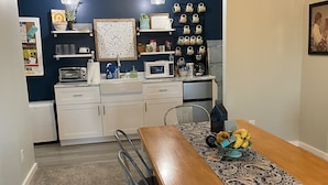 A compact kitchen with white cabinets, a microwave, and a coffee maker on the counter. There are two pendant lights hanging above the counter, and a patterned table runner on the dining table.