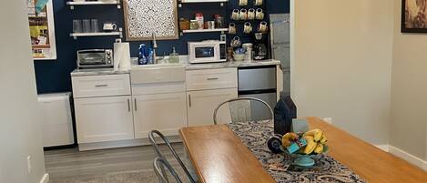 A compact kitchen with white cabinets, a microwave, and a coffee maker on the counter. There are two pendant lights hanging above the counter, and a patterned table runner on the dining table.