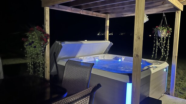 An outdoor hot tub with a wooden pergola structure, surrounded by chairs and a hanging planter.
