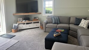 A living room with a flat-screen TV mounted on a wooden shelf, a grey sofa with patterned cushions, a blue ottoman, and a window with a view of greenery.