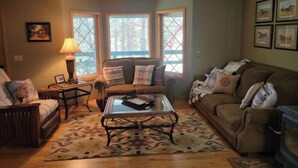 A cozy living room with a sofa, armchair, and a coffee table. There are windows with a view of trees and a fireplace. - Secluded Montana home on five acres (Rexford)