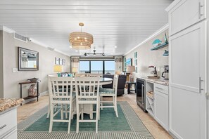 Une cuisine avec des meubles blancs, un coin repas avec une table et des chaises, un lustre et une vue sur l’extérieur.