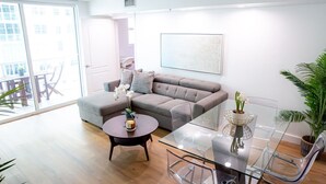 A modern living room with a grey sectional sofa, a glass coffee table, and a glass dining table with chairs. There is a potted plant and a small table with a candle and flowers. - Awesome 4BR APT @Brickell, Free Parking (Miami)