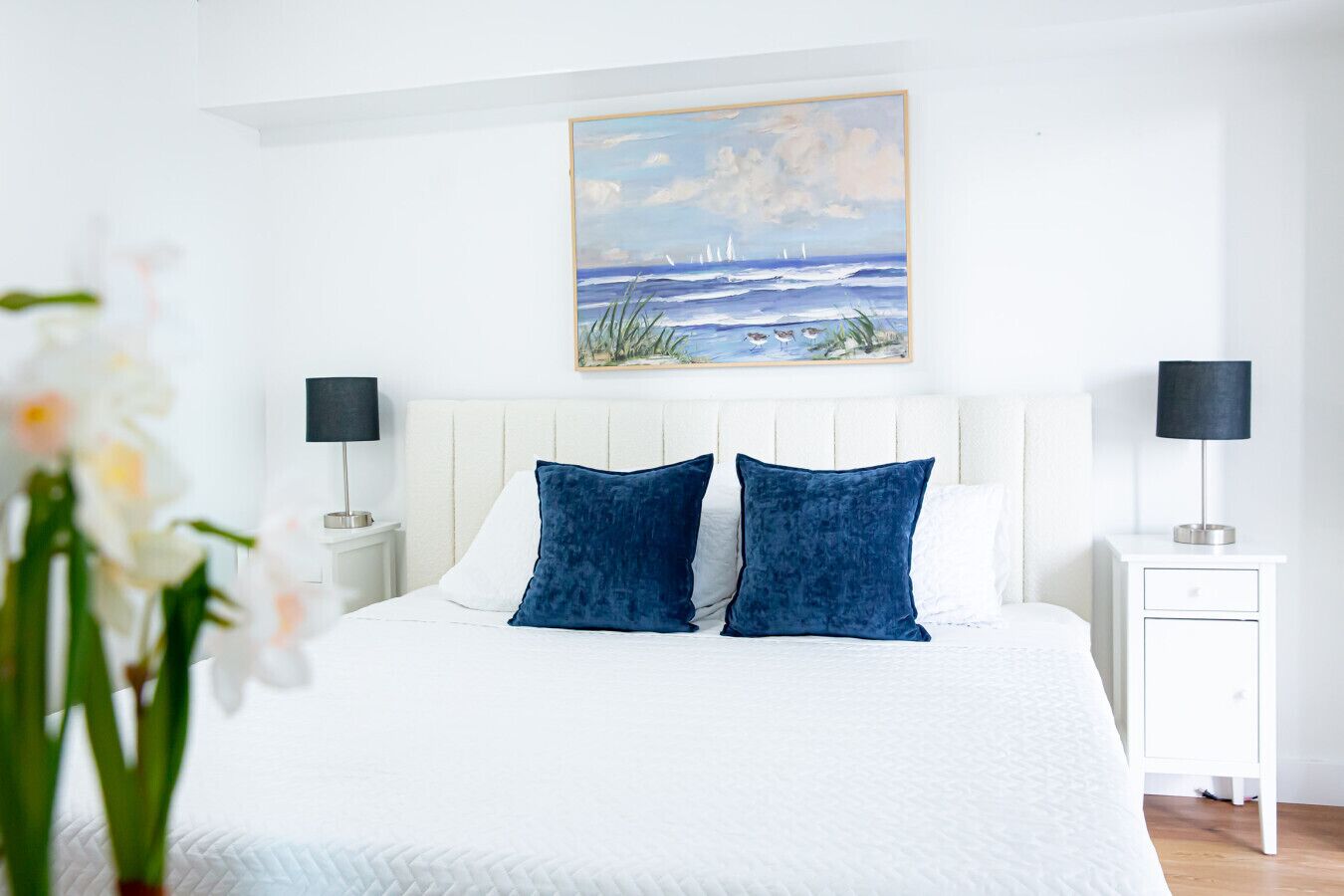 A bedroom with a white bed, two blue pillows, a white headboard, a nightstand with a lamp, and a painting of a seascape above the bed.