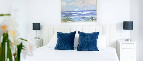 A bedroom with a white bed, two blue pillows, a white headboard, a nightstand with a lamp, and a painting of a seascape above the bed.