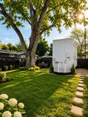 Un jardin avec un grand arbre, un abri de jardin blanc et une allée pavée.