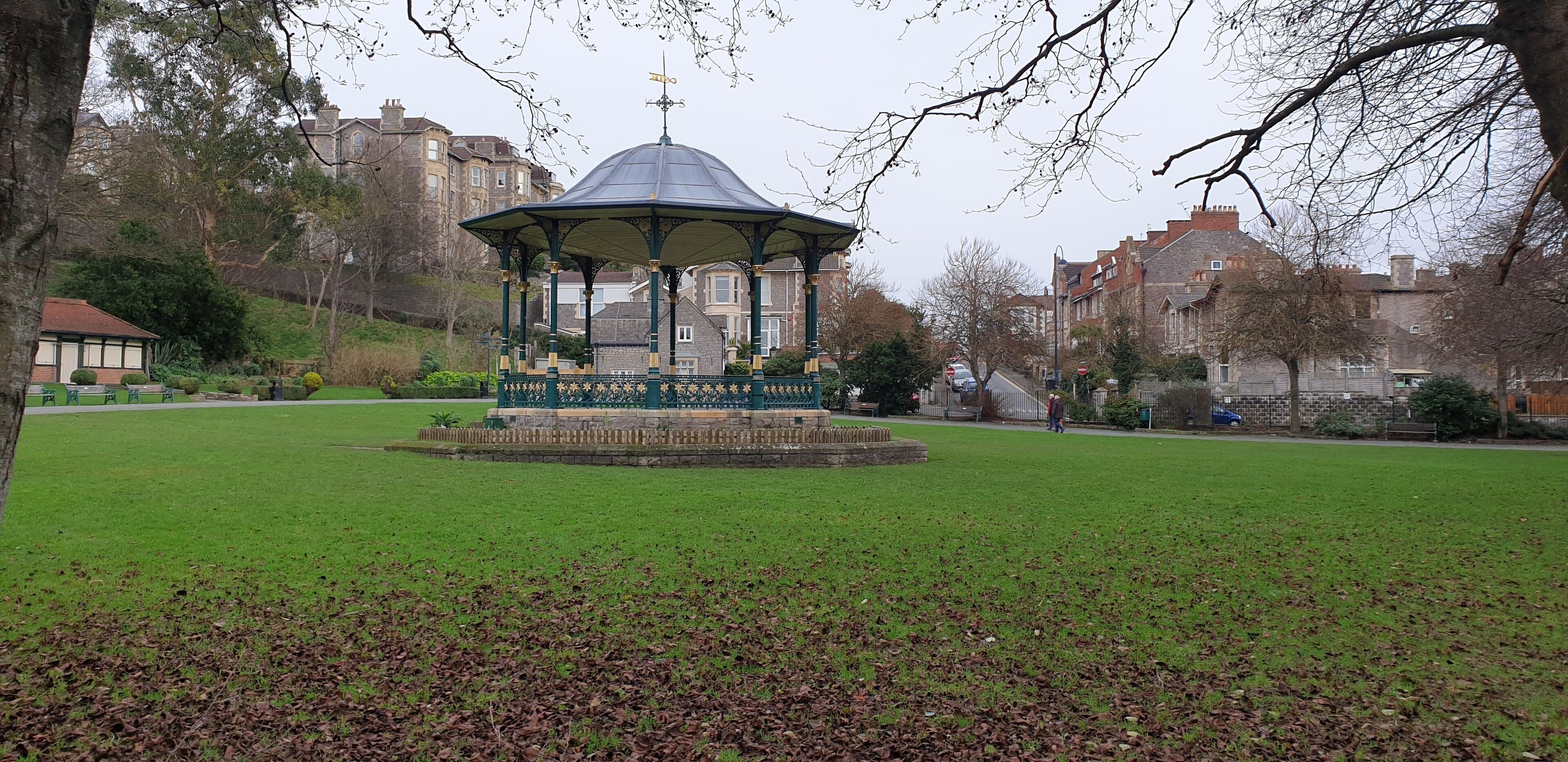 Un gazebo in un parco con una fontana al centro, circondato da erba e alberi.