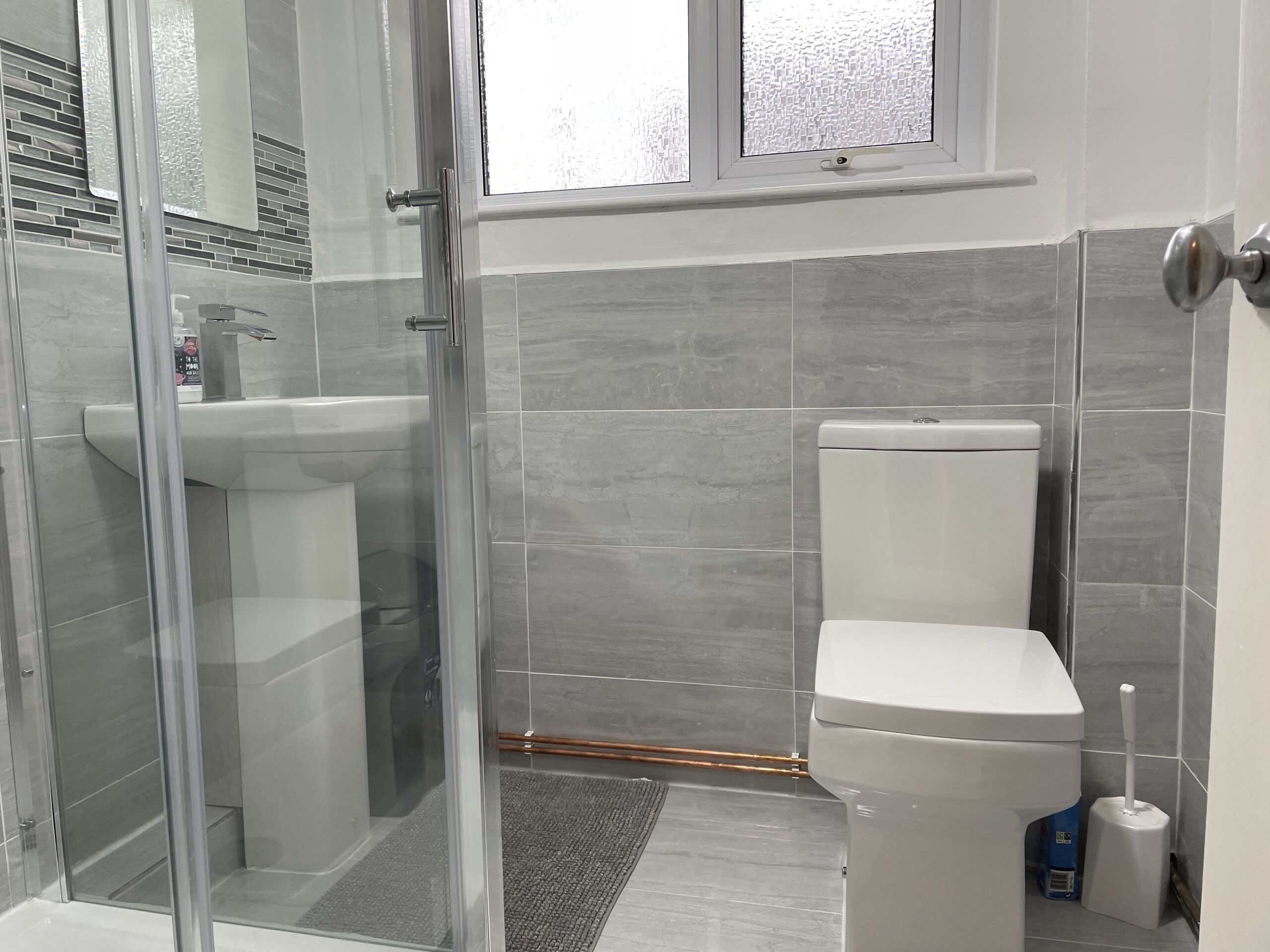 Et moderne badeværelse med en brusekabine i glas, et hvidt toilet og vægge belagt med grå fliser.