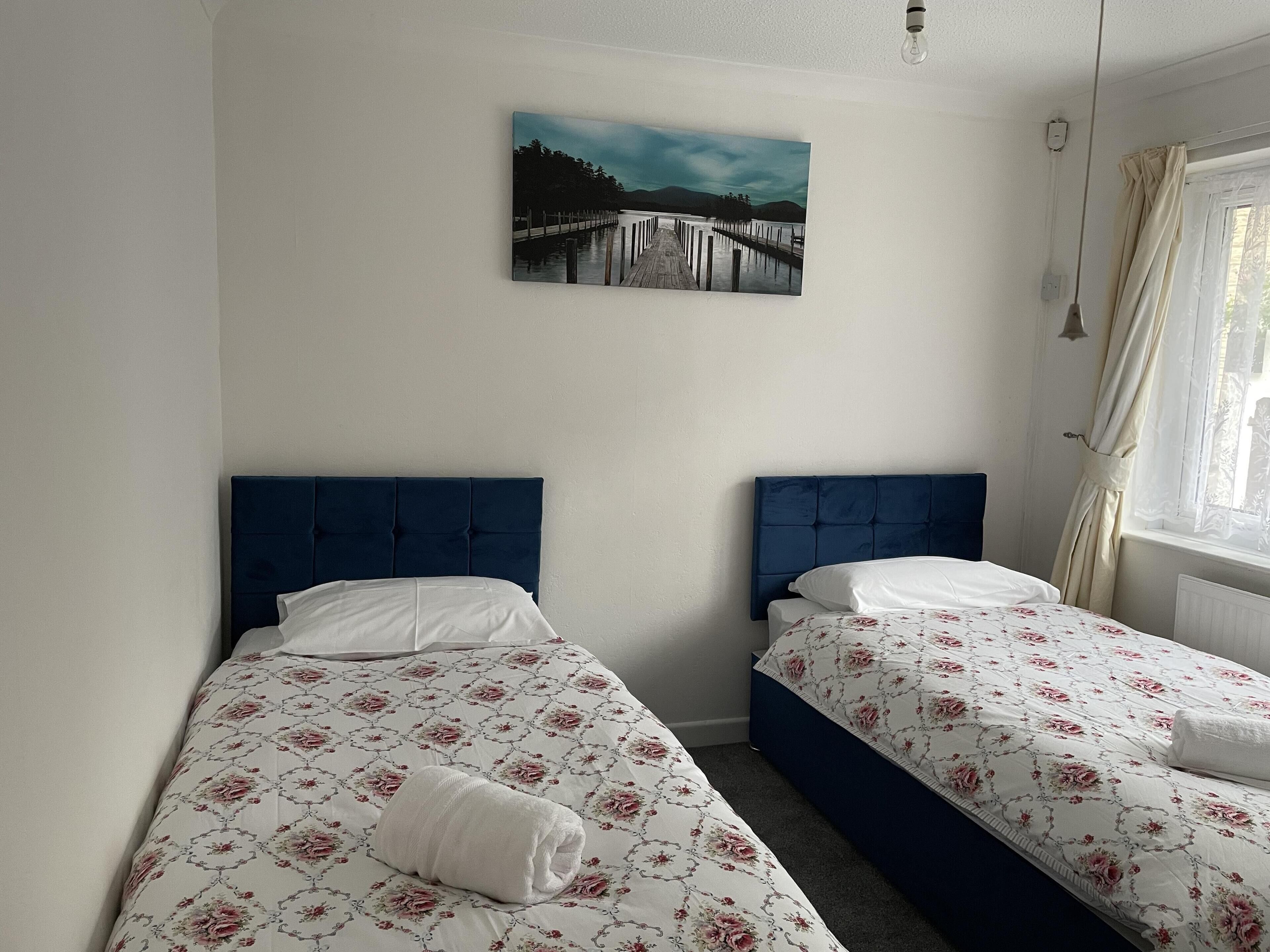 A hotel room with two beds, each with a blue headboard and floral bedding. A painting of a pier and mountains is on the wall.