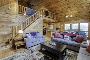 A wooden cabin interior with two sofas, a coffee table, a staircase, and a kitchen area.