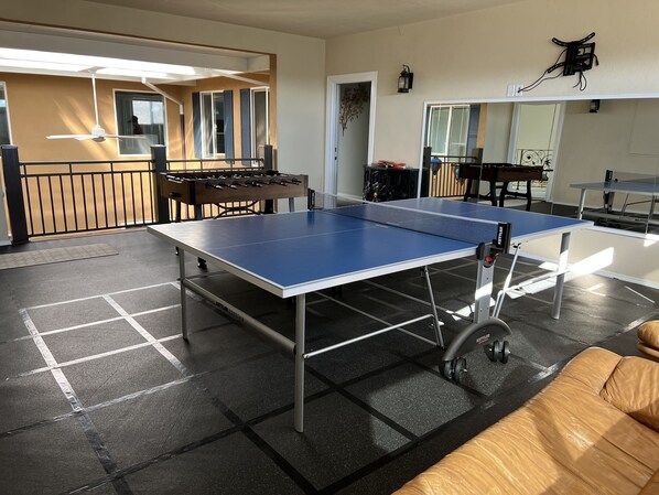 Ett bordtennisbord i ett rum med ett fotbollsspel, en soffa och ett köksområde som syns genom en öppen dörr.