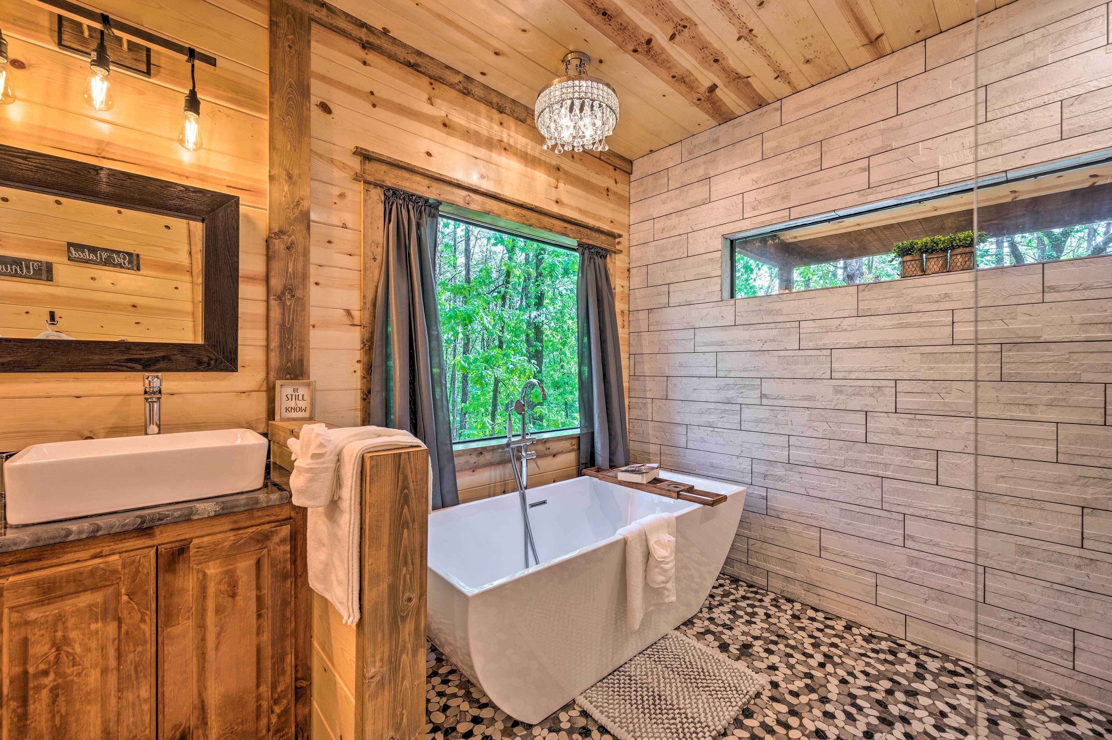 Une salle de bain rustique, avec une cabine en bois, une grande baignoire, un meuble-lavabo en bois et une fenêtre donnant sur un paysage verdoyant.
