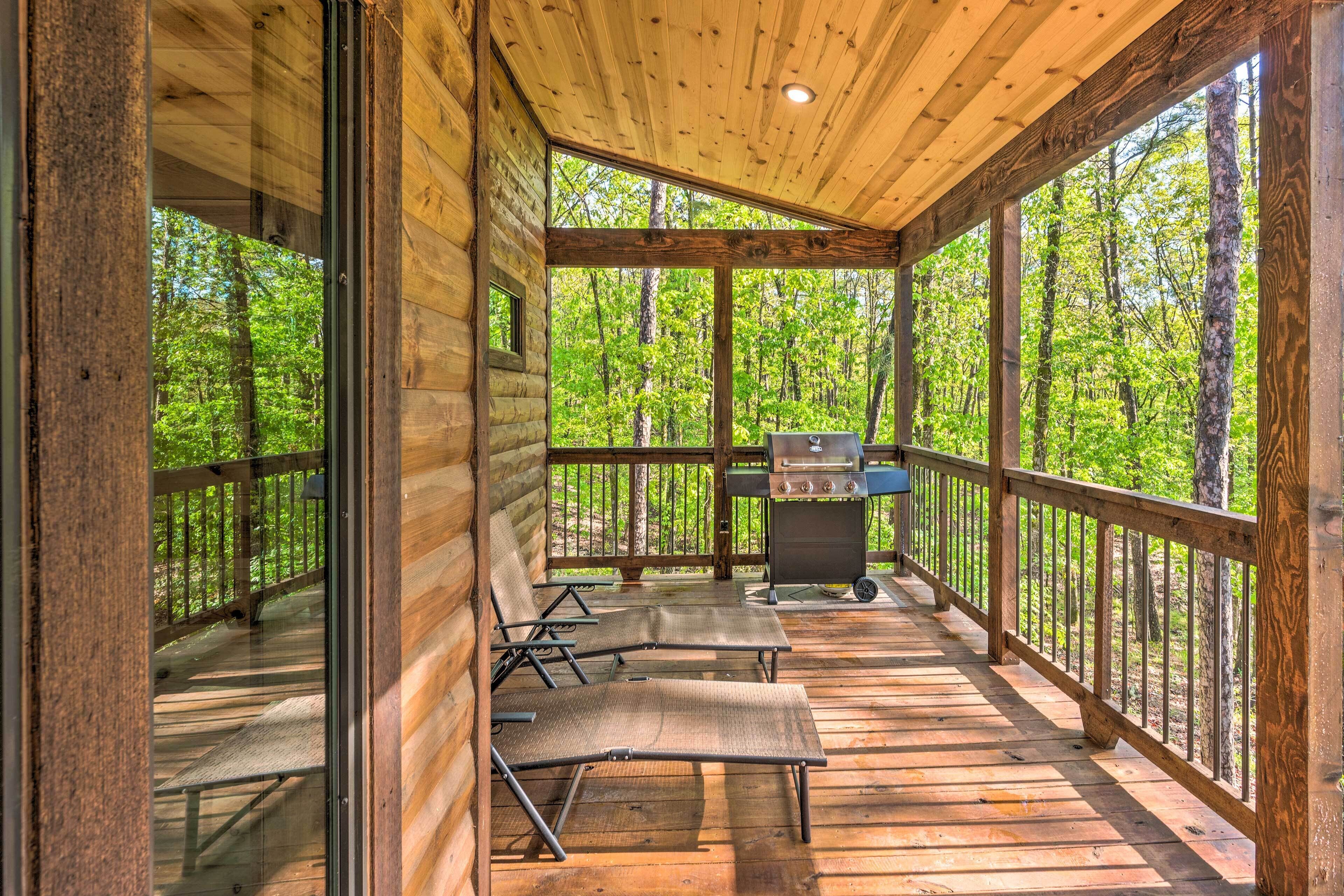 Une terrasse en bois couverte, dotée d’un barbecue et de chaises longues, entourée d’une forêt.