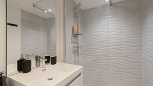 Een moderne badkamer met een grote spiegel, een witte wastafel, twee zwarte zeepdispensers, een handdouche en geribbelde wandtegels.