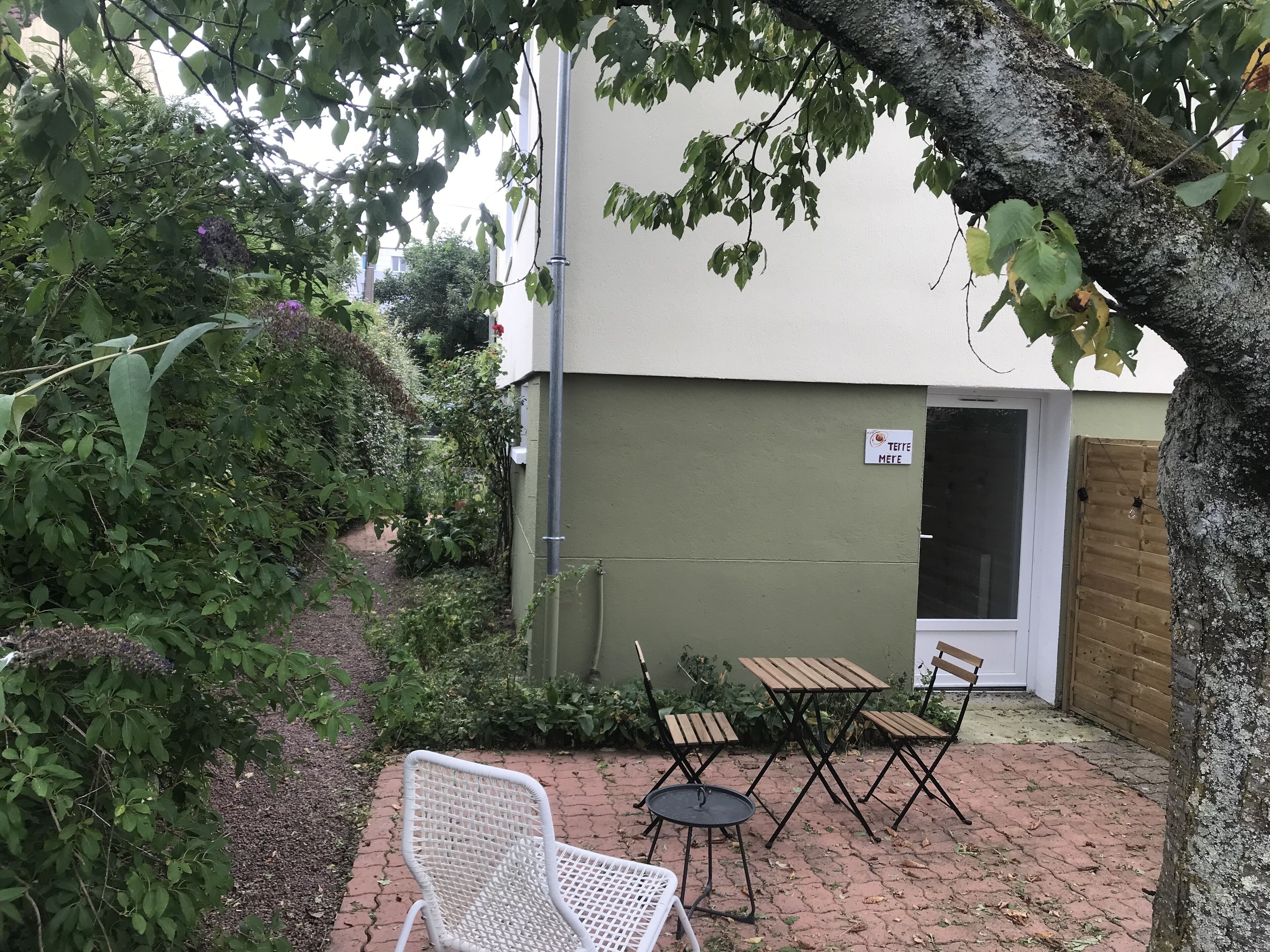 Un espace patio comprenant une chaise en osier blanc, deux tables en bois et quatre chaises. Un panneau avec un logo est visible sur le mur.